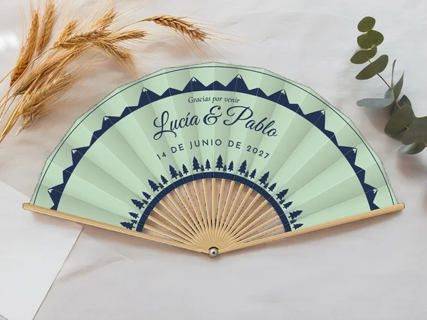 Abanico de boda personalizado “Pinos y montañas” con los nombres Lucía & Pablo, crestas de montañas en el borde y fila de abetos abajo, tela color verde agua e impresión en azul marino, sobre una mesa de boda