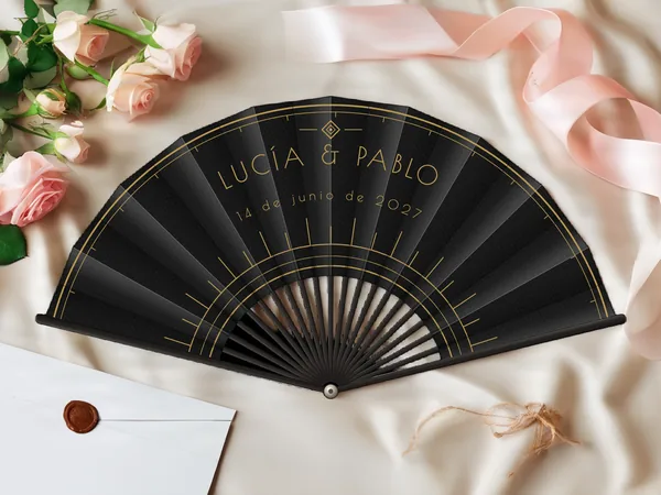 Abanico de boda personalizado “Art déco” con los nombres Lucía & Pablo, arcos finos y rayos gráficos art déco alrededor del centro, tela color negro e impresión en oro, sobre una mesa de boda