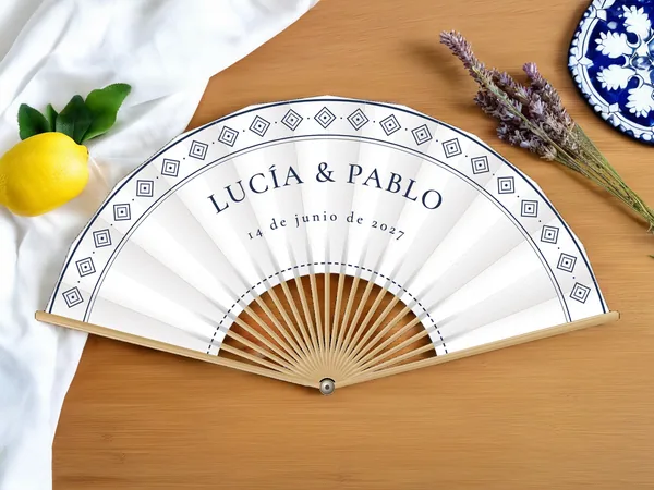 Abanico de boda personalizado “Talavera” con los nombres Lucía & Pablo, friso de azulejos en rombo a lo largo del borde, tela color blanco e impresión en azul marino, sobre una mesa de boda