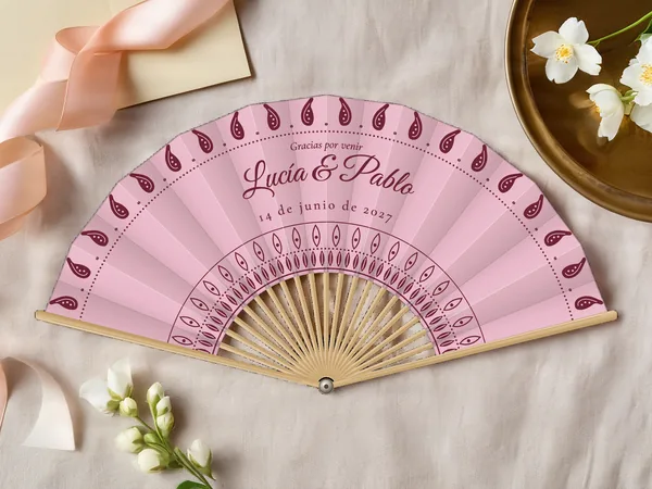Abanico de boda personalizado “Paisley” con los nombres Lucía & Pablo, gotas de cachemir a lo largo del borde y rosetón alrededor del centro, tela color palo de rosa e impresión en vino, sobre una mesa de boda