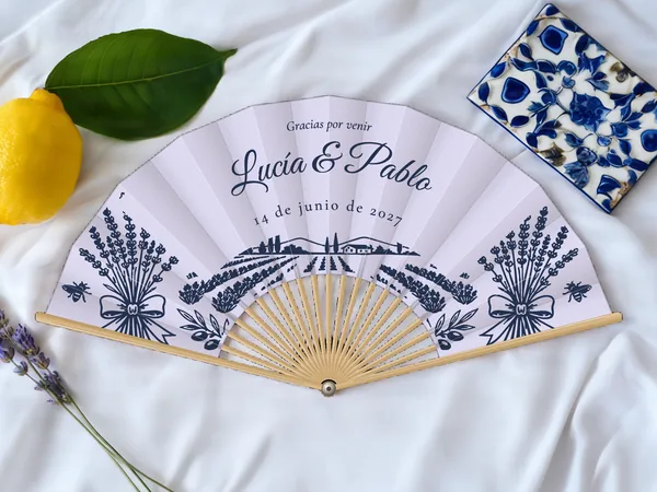 Abanico de boda personalizado “Campo de lavanda”, ramos de lavanda, abejas y campo de lavanda bajo los nombres Lucía & Pablo, tela color lavanda e impresión en azul marino, sobre una mesa de boda