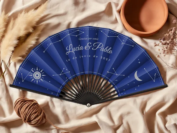 Abanico de boda personalizado “Celeste”, constelaciones, sol y luna creciente esparcidos alrededor de los nombres Lucía & Pablo, tela color azul marino e impresión en plata, sobre una mesa de boda