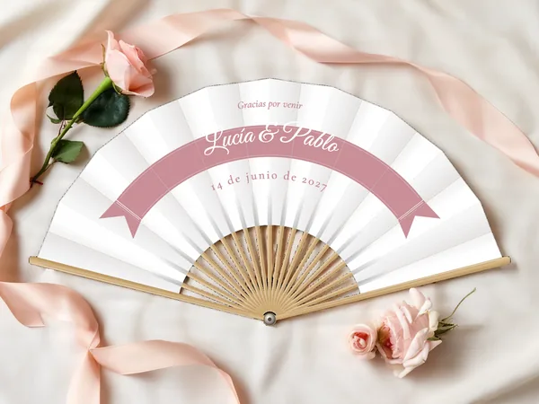 Abanico de boda personalizado “Listón”, ancha cinta en arco que lleva los nombres Lucía & Pablo, tela color blanco e impresión en palo de rosa, sobre una mesa de boda