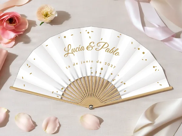 Abanico de boda personalizado “Confeti”, pequeños confetis esparcidos alrededor de los nombres Lucía & Pablo, tela color blanco e impresión en oro, sobre una mesa de boda