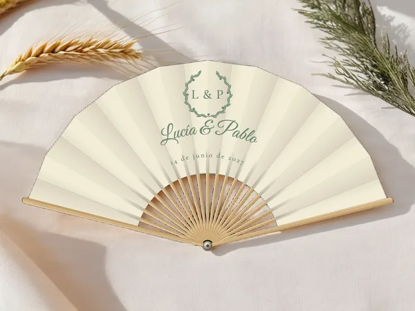 Abanico de boda personalizado “Corona de olivo”, corona de follaje rodeando las iniciales sobre los nombres Lucía & Pablo, tela color marfil e impresión en verde salvia, sobre una mesa de boda