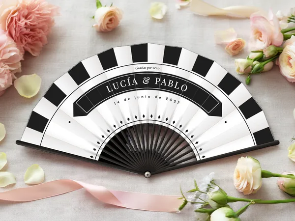 Abanico de boda personalizado “De etiqueta”, rayas en damero en el borde y banda bajo los nombres Lucía & Pablo, tela color blanco e impresión en negro, sobre una mesa de boda