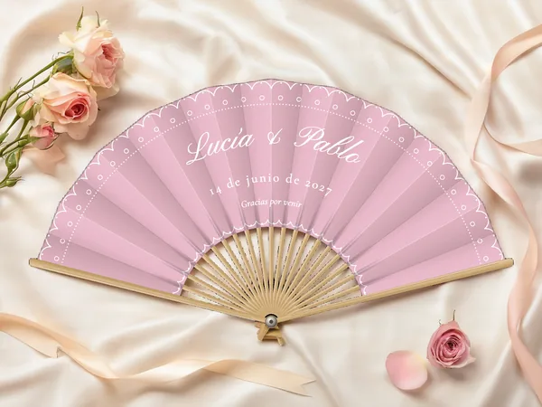 Abanico de boda personalizado “Encaje” con los nombres Lucía & Pablo, borde festoneado tipo encaje y punteado siguiendo el contorno, tela color palo de rosa e impresión en blanco, sobre una mesa de boda