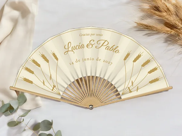 Abanico de boda personalizado “Espigas de trigo” con los nombres Lucía & Pablo, espigas de trigo erguidas a cada lado y filete en el borde, tela color marfil e impresión en oro, sobre una mesa de boda