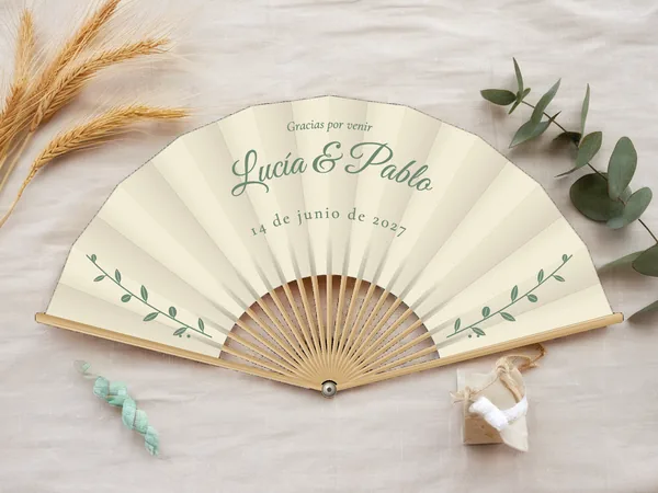 Abanico de boda personalizado “Eucalipto”, finas ramas con hojas a cada lado bajo los nombres Lucía & Pablo, tela color marfil e impresión en verde salvia, sobre una mesa de boda