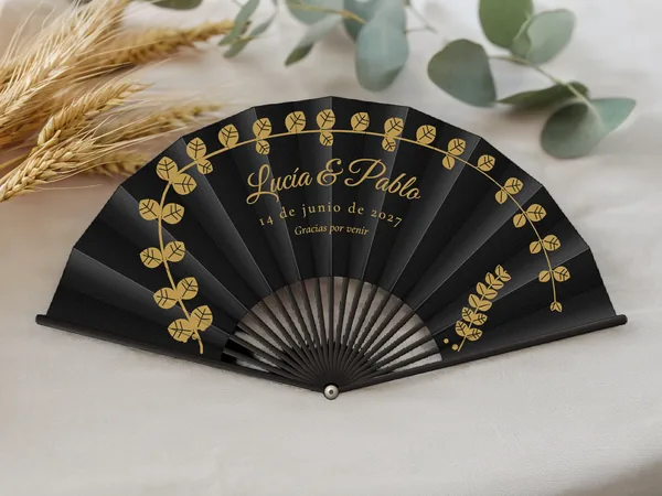 Abanico de boda personalizado “Eucalipto dorado”, ramas de eucalipto en arco enmarcando los nombres Lucía & Pablo, tela color negro e impresión en oro, sobre una mesa de boda
