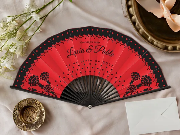 Abanico de boda personalizado “Flamenco” con los nombres Lucía & Pablo, volante de encaje con lunares en el borde y claveles en las esquinas, tela color rojo e impresión en negro, sobre una mesa de boda