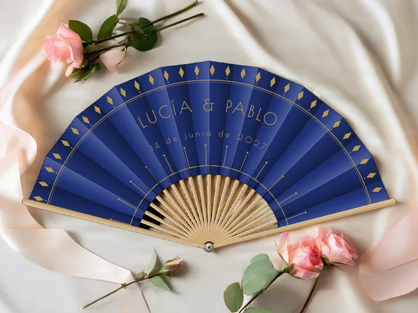 Abanico de boda personalizado “Gatsby”, rombos a lo largo del borde y rayos art déco bajo los nombres Lucía & Pablo, tela color azul marino e impresión en oro, sobre una mesa de boda