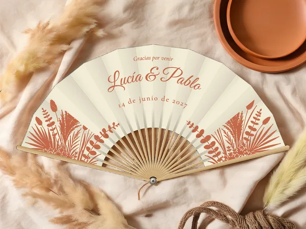 Abanico de boda personalizado “Pasto de pampa” con los nombres Lucía & Pablo, hierba de la pampa, palmas y follaje seco en las esquinas inferiores, tela color marfil e impresión en terracota, sobre una mesa de boda
