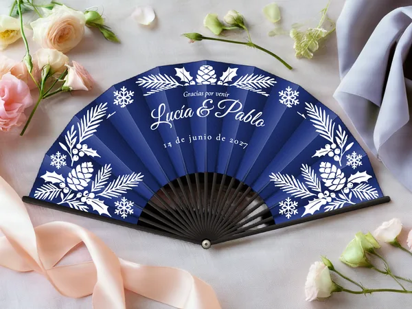 Abanico de boda personalizado “Invierno escarchado”, ramas de abeto, piñas, acebo y copos enmarcando los nombres Lucía & Pablo, tela color azul marino e impresión en blanco, sobre una mesa de boda