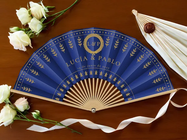 Abanico de boda personalizado “Laurel real”, corona de laurel sobre los nombres Lucía & Pablo y ramitas en hileras, tela color azul marino e impresión en oro, sobre una mesa de boda