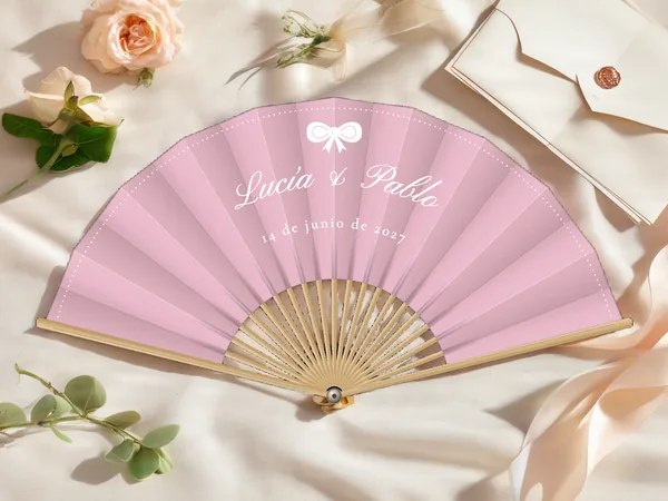 Abanico de boda personalizado “Moño”, pequeño lazo sobre los nombres Lucía & Pablo y punteado a lo largo del borde, tela color palo de rosa e impresión en blanco, sobre una mesa de boda