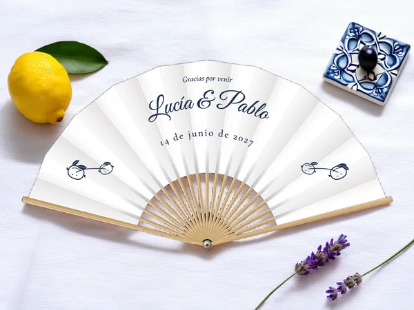 Abanico de boda personalizado “Limonero”, pequeña rama con dos limones a cada lado bajo los nombres Lucía & Pablo, tela color blanco e impresión en azul marino, sobre una mesa de boda