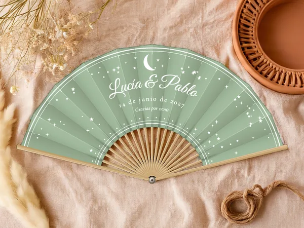 Abanico de boda personalizado “Luna y estrellas”, luna creciente y estrellas esparcidas alrededor de los nombres Lucía & Pablo, tela color verde salvia e impresión en blanco, sobre una mesa de boda