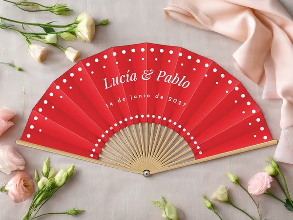 Abanico de boda personalizado “Lunares” con los nombres Lucía & Pablo, hileras de lunares a lo largo del borde y alrededor del centro, tela color rojo e impresión en blanco, sobre una mesa de boda