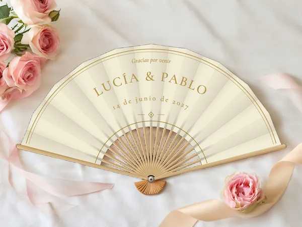 Abanico de boda personalizado “Marfil y oro”, doble trazo fino siguiendo el borde y pequeño rombo bajo los nombres Lucía & Pablo, tela color marfil e impresión en oro, sobre una mesa de boda