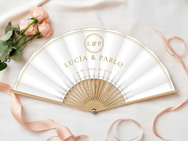Abanico de boda personalizado “Monograma dorado”, medallón con iniciales sobre los nombres Lucía & Pablo y doble trazo en el borde, tela color blanco e impresión en oro, sobre una mesa de boda