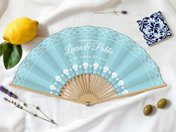 Abanico de boda personalizado “Frente al mar”, olas en el borde y arco de conchas bajo los nombres Lucía & Pablo, tela color azul cielo e impresión en blanco, sobre una mesa de boda