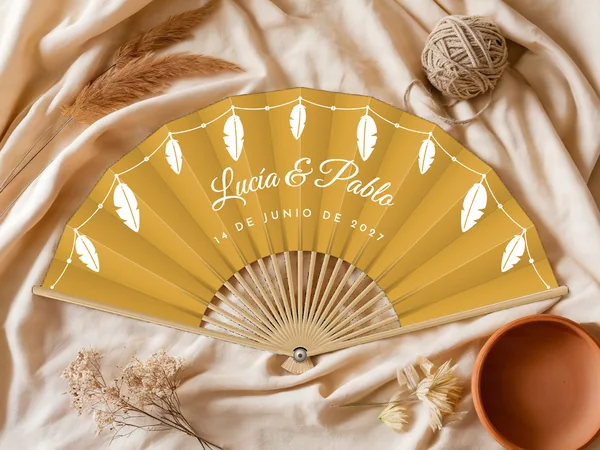 Abanico de boda personalizado “Plumas” con los nombres Lucía & Pablo, plumas colgando de una guirnalda de perlas por el borde, tela color mostaza e impresión en blanco, sobre una mesa de boda