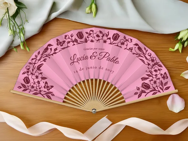 Abanico de boda personalizado “Primavera en flor”, guirnalda de flores, pájaros y mariposas enmarcando los nombres Lucía & Pablo, tela color rosa e impresión en vino, sobre una mesa de boda