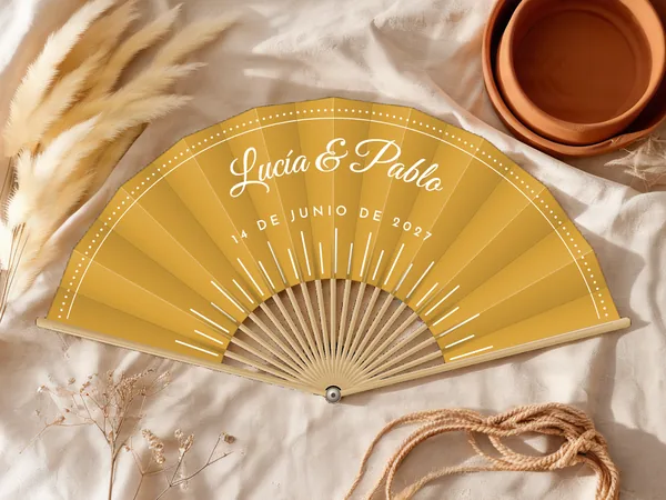 Abanico de boda personalizado “Rayos de sol” con los nombres Lucía & Pablo, rayos finos desde abajo y friso punteado en el borde, tela color mostaza e impresión en blanco, sobre una mesa de boda
