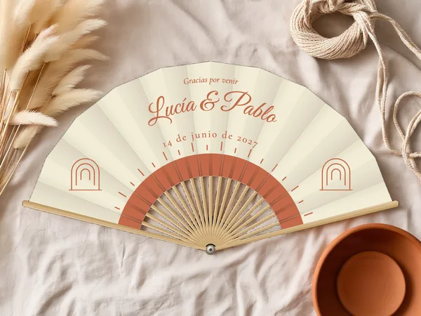 Abanico de boda personalizado “Sol boho”, medio sol radiante bajo los nombres Lucía & Pablo y pequeños arcos a los lados, tela color marfil e impresión en terracota, sobre una mesa de boda