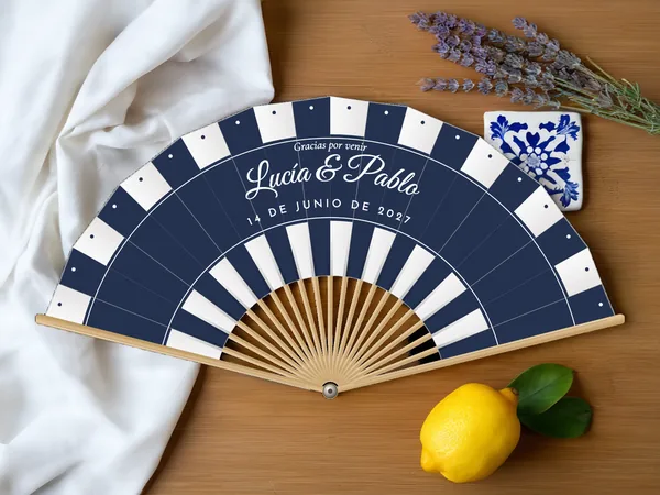 Abanico de boda personalizado “Sombrilla” con los nombres Lucía & Pablo, rayas de sombrilla en bandas por el borde y abajo, tela color blanco e impresión en azul marino, sobre una mesa de boda
