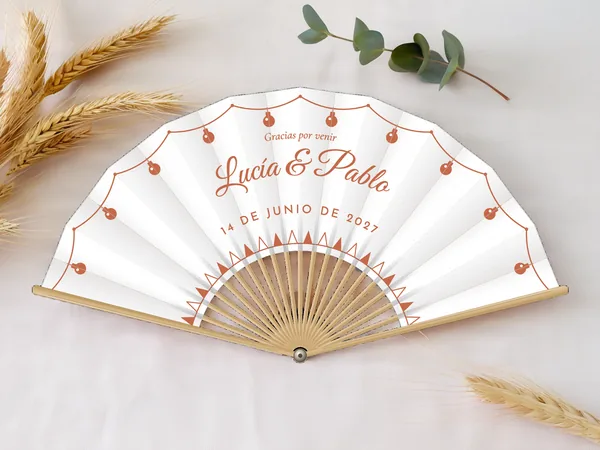 Abanico de boda personalizado “Kermés” con los nombres Lucía & Pablo, guirnalda de bombillas por el borde y friso de banderines abajo, tela color blanco e impresión en terracota, sobre una mesa de boda